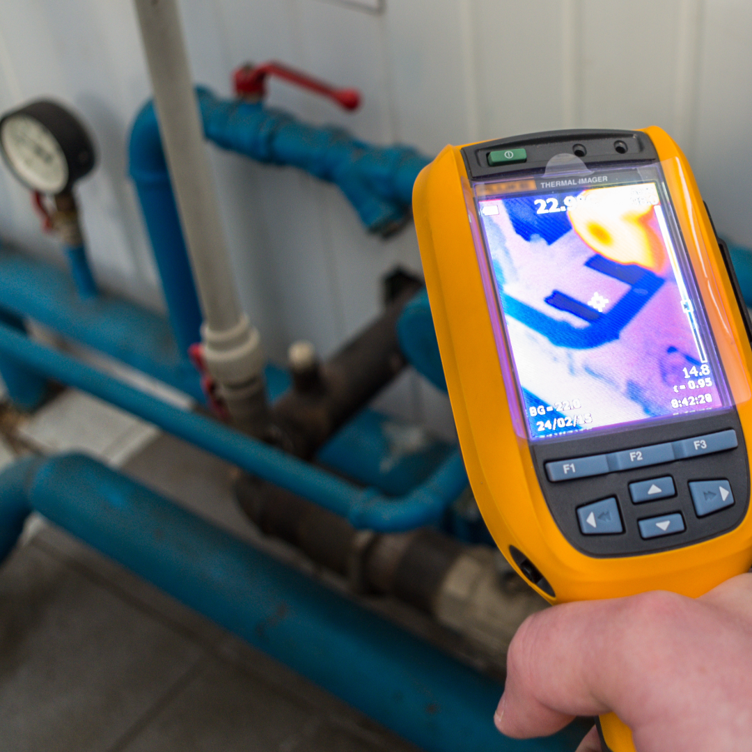 The image shows a thermal imaging device being used to inspect a system of pipes and valves. The screen on the device displays a thermal heat map of the area, which likely indicates temperature variations in the pipes. Thermal imaging is commonly used in energy audits or maintenance inspections to identify heat loss, leaks, or other inefficiencies in systems such as heating, ventilation, and air conditioning (HVAC), electrical systems, or industrial machinery.